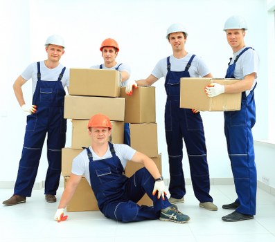 A group of five professional movers in Ealing, dressed in blue overalls, white shirts, and safety helmets, pose for a photo inside a bright residential space with white walls and large windows. Three of them stand behind and beside a stack of cardboard boxes, each holding or supporting a box, demonstrating teamwork during a house removal. One mover is kneeling in front, with one knee on the floor, and is wearing an orange safety helmet, smiling towards the camera. The other four, wearing white safety helmets, are positioned to the sides, with some resting hands on the boxes or holding them securely. The team appears prepared for a home relocation, with the boxes being of various sizes, some sealed with packing tape, and others open showing contents like books or small household items. The environment suggests a clean, well-lit room, likely part of a residential property in the Ealing area, highlighting the professionalism and readiness of Ealing Man with Van's house removal services in West London.