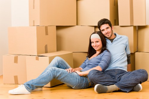 A young man and woman with light skin are sitting on the wooden floor of a residential space, surrounded by stacked large cardboard moving boxes in various sizes. The woman, with long dark hair and wearing a white headband and casual blue jeans and top, is leaning back slightly and smiling at the camera. The man, with short dark hair and dressed in a light blue shirt and dark trousers, is sitting behind her with one arm resting on her shoulder, also smiling. The background shows a neutral-colored wall, suggesting they are inside a house or flat that is in the process of being packed or moved. The scene depicts a moment of preparation for house removal or relocation, aligning with professional services offered by Ealing Man with Van in the Ealing area, with a focus on efficient and approachable removals. The environment is well-lit with natural or soft indoor lighting, highlighting the textures of the boxes and the casual clothing of the individuals, emphasizing a friendly, organized moving process typical of local house removal services in Ealing, West London.