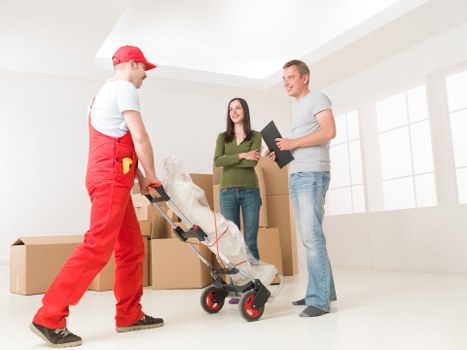 In a bright, minimally decorated room with white walls and large windows allowing natural light, a house removal process is underway. A man dressed in red work overalls, with a cap, is handling a trolley loaded with a wrapped household item, likely a fragile object, positioned in the foreground on the left side. Behind him, several large cardboard boxes are stacked, some open and others sealed, indicating packing activity. In the background, a woman with long dark hair, wearing a green sweater and blue jeans, is holding a clipboard and engaging in conversation with a man in casual grey t-shirt and jeans, who is smiling and holding some papers. The scene suggests a professional house removal or relocation service, with the team coordinating and preparing for moving household belongings. The setting appears to be an interior of a house, possibly a living room or hallway, with ample natural daylight streaming in, emphasizing a professional, organized, and calm atmosphere typical of Ealing-based removals services.