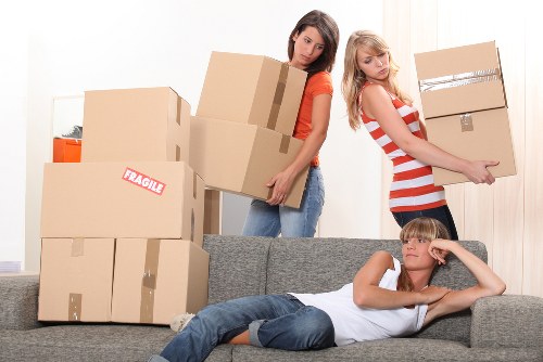 In a residential living room, two young women are engaged in a house removal activity. One woman, wearing a red top and jeans, is holding a medium-sized cardboard box with brown tape sealing the top edges, positioned behind a stack of various-sized boxes, some with labels and orange storage containers on top. She stands on the floor near a grey fabric sofa, which is partially visible in the foreground, and appears to be preparing to load or move the boxes. The other woman, dressed in a red and white striped top and dark pants, is carrying a large cardboard box with packing tape and cardboard reinforcements visible on the edges, and is walking away from the sofa toward the door, suggesting she is either transitioning to another room or preparing to load the boxes into a vehicle. A young boy is lying on the sofa in a relaxed pose, resting his head on one hand, wearing a white T-shirt and blue jeans, with short blond hair. The background displays a light-colored wall and a curtain covering a window, illuminated by natural daylight, creating a neutral, well-lit environment appropriate for a house removal setting, with subtle hints of a suburban residential area near Ealing in the background.