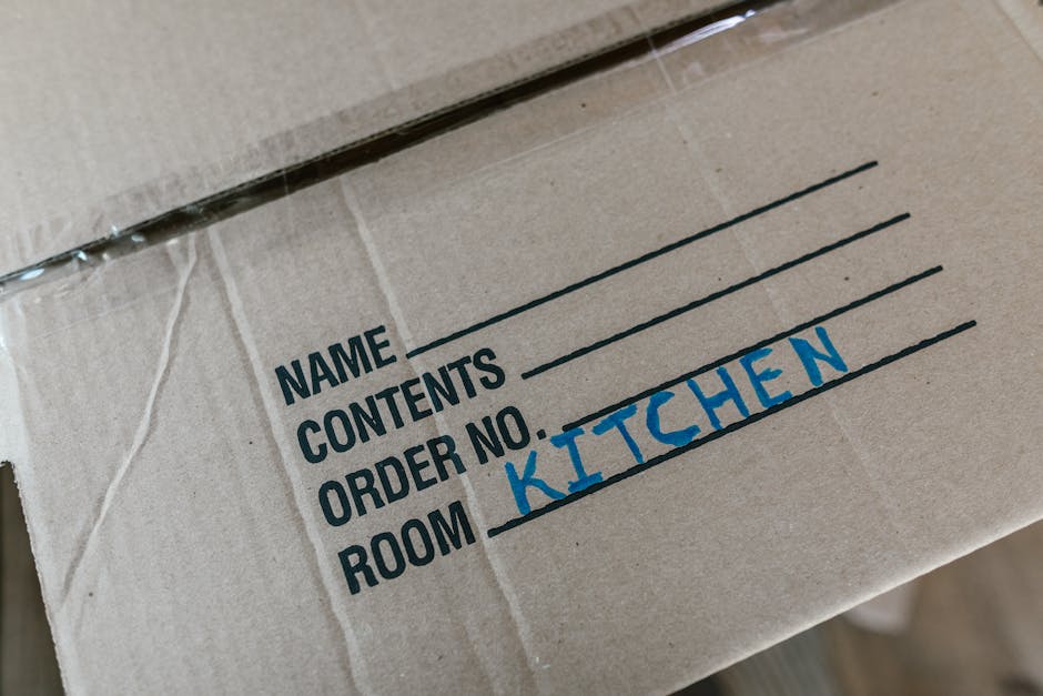 Close-up of a cardboard moving box with a shipping label visible, indicating the contents and destination. The label shows the room name 'KITCHEN' handwritten in blue marker, with predefined fields fo